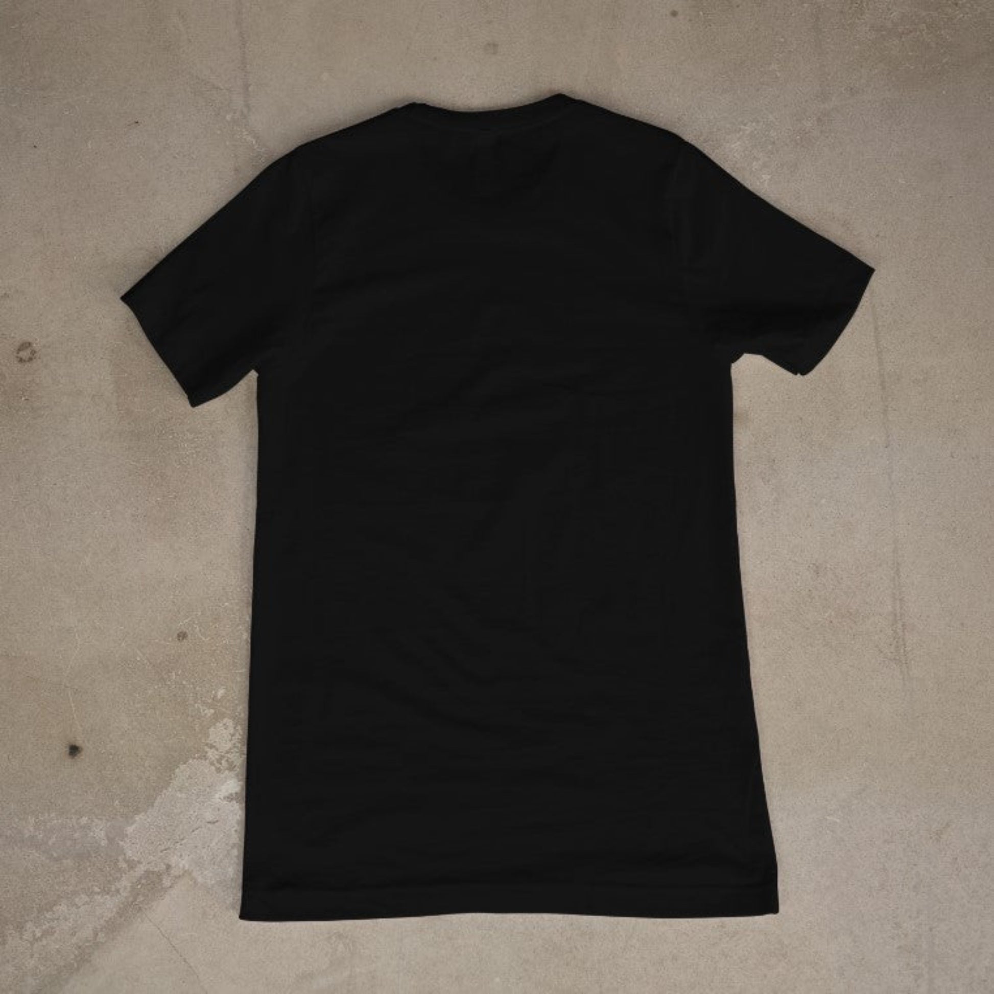 "Back view of a plain black unisex cotton blend t-shirt laid flat on a textured concrete background"