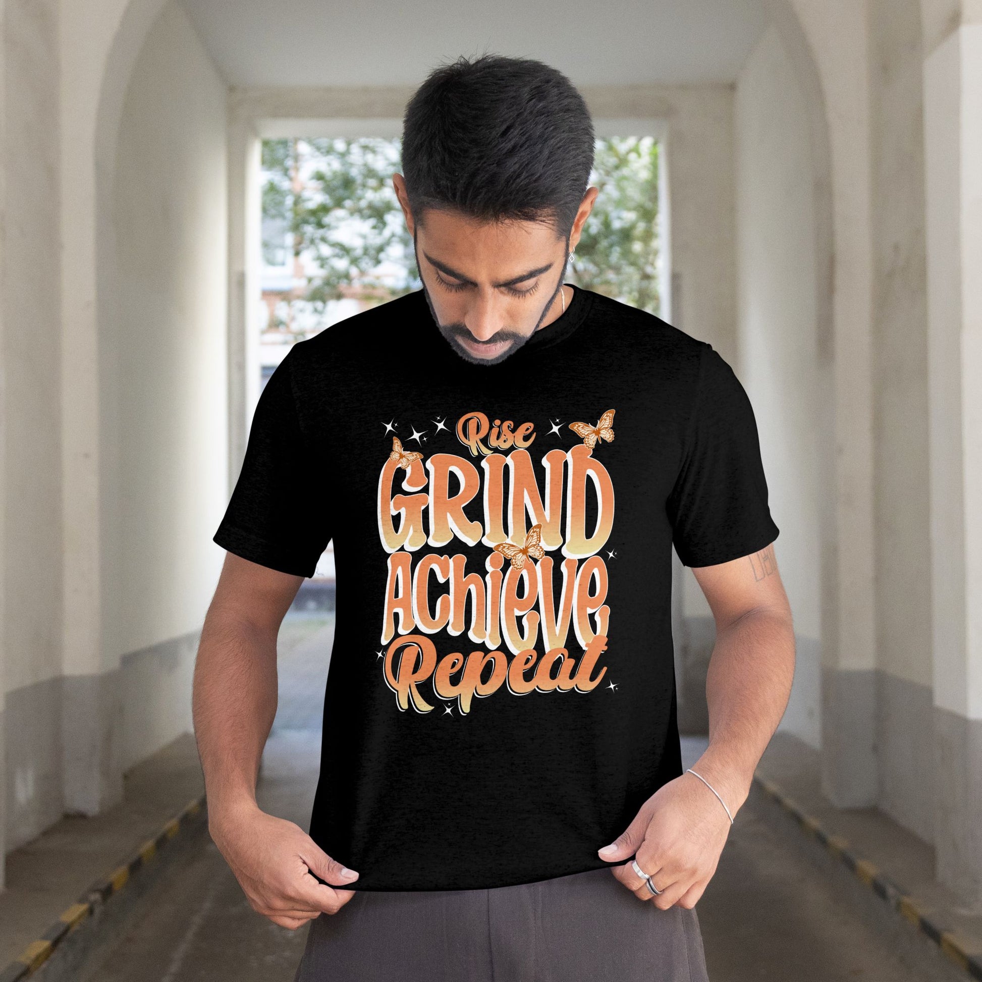 Man wearing a black t-shirt with 'Rise Grind Achieve Repeat' text in an urban setting