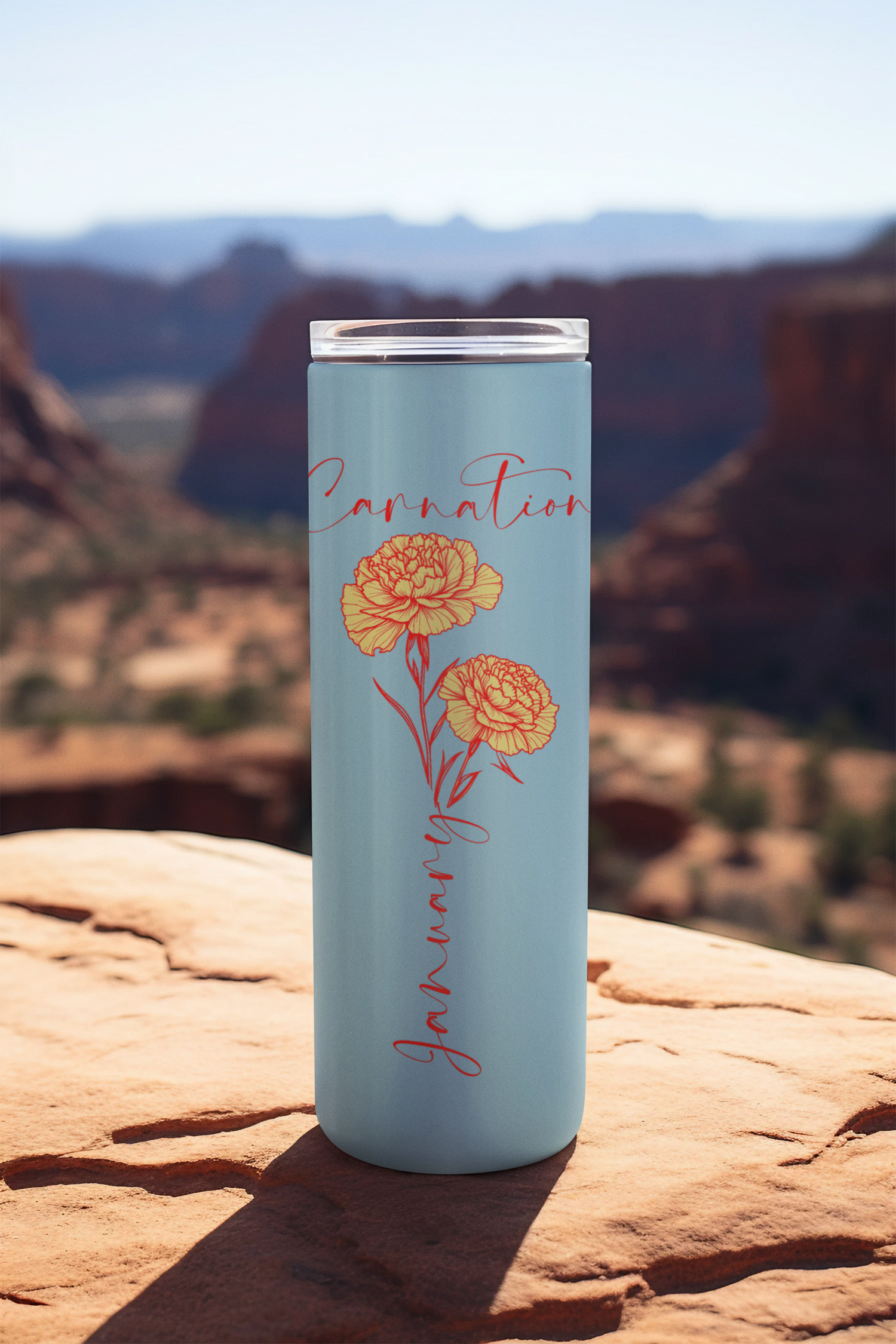 20oz stainless steel tumbler with a light blue background featuring the word “January” and illustrated carnations, photographed outdoors on a scenic red rock landscape.