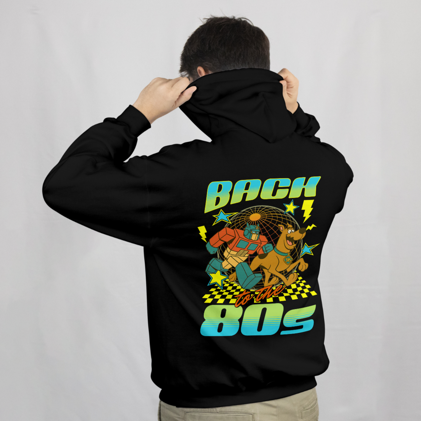 Man wearing black hoodie with large back graphic featuring retro 80s design, bold text reading “Back to the 80s,” cartoon-style robot and dog, stars, lightning bolts, and neon colors