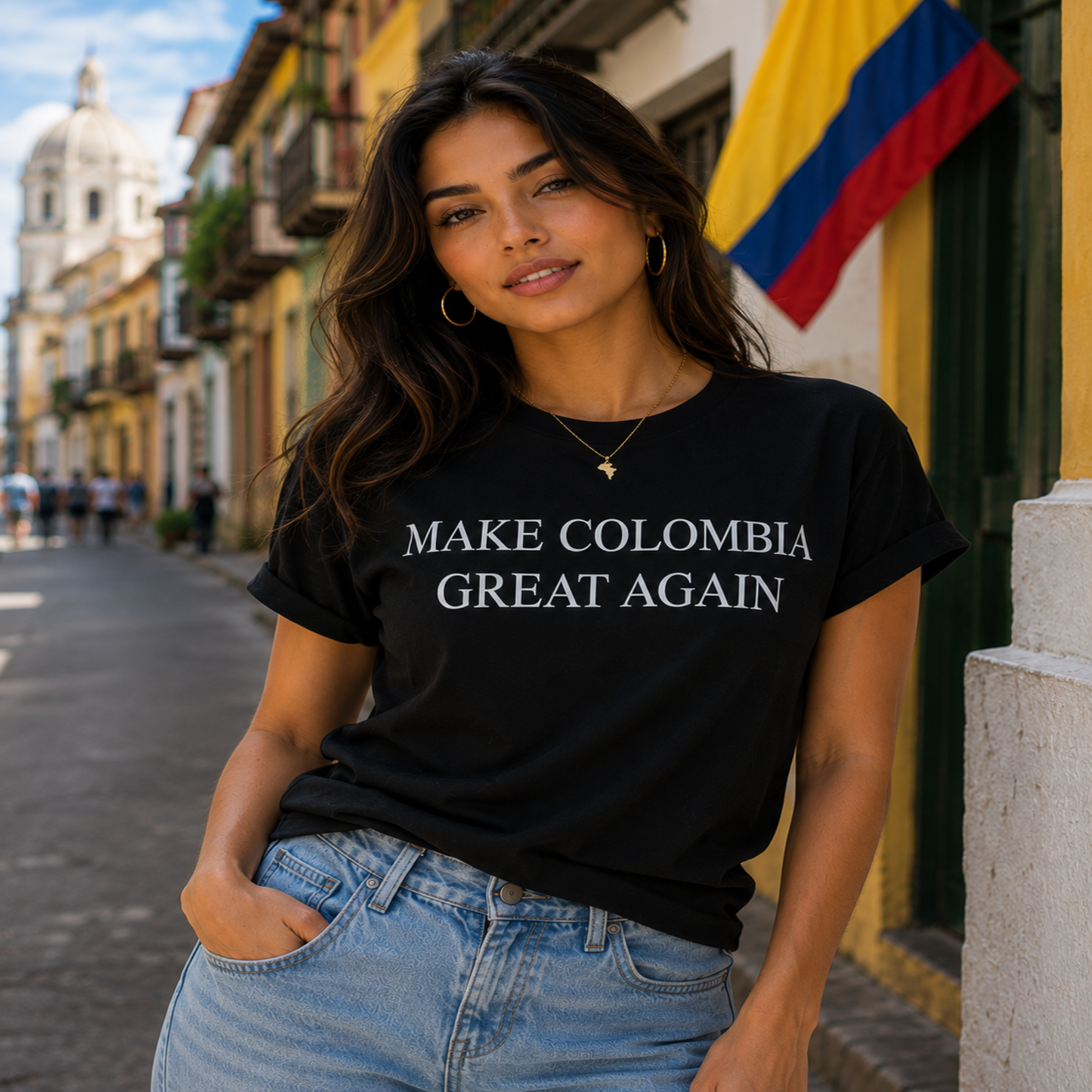 A woman wearing a black graphic t-shirt with the text 'MAKE COLOMBIA GREAT AGAIN' in white serif letters, paired with light blue jeans, standing on a street with a Colombian flag in the background.