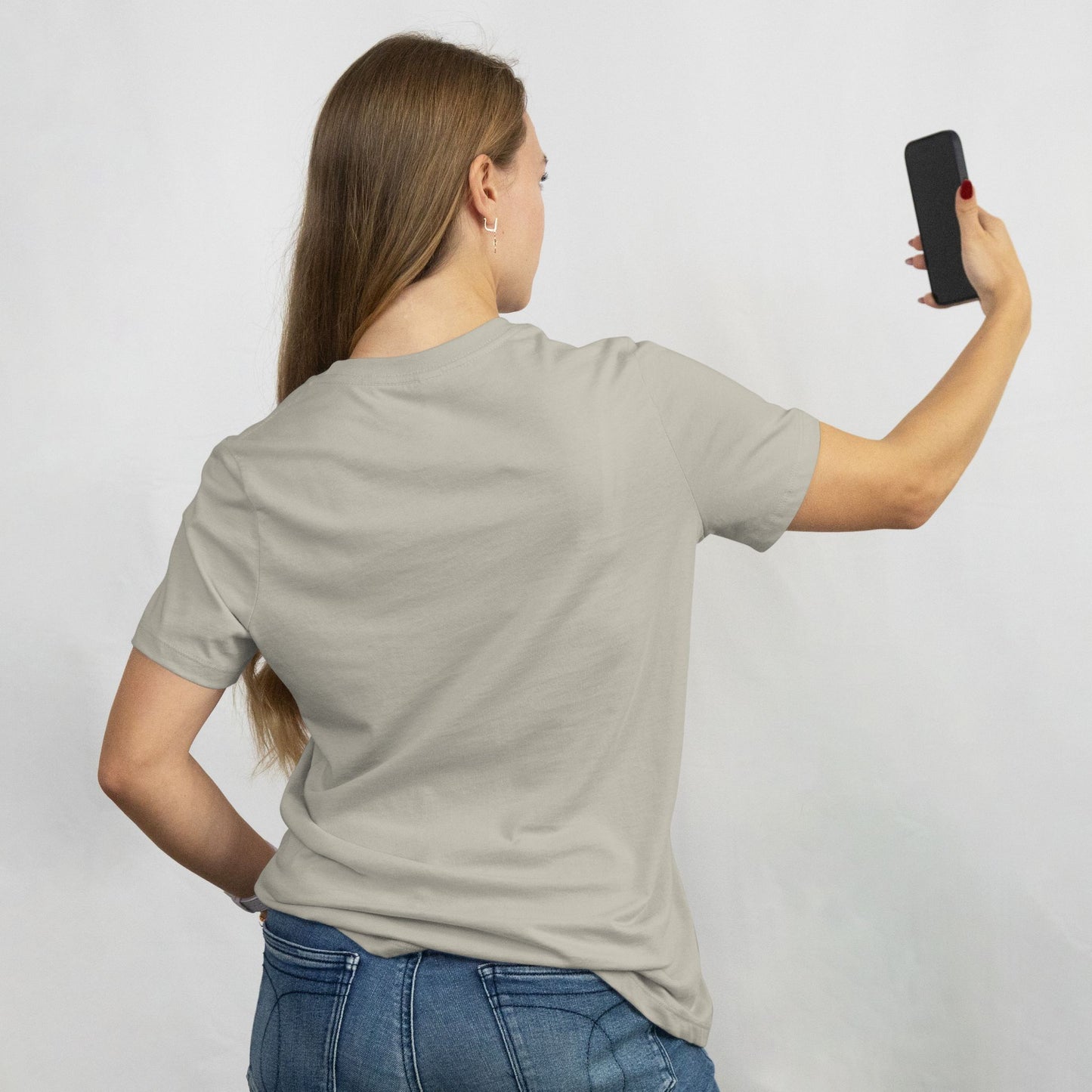 Woman taking a selfie wearing a plain gray t-shirt and blue jeans on a light gray background