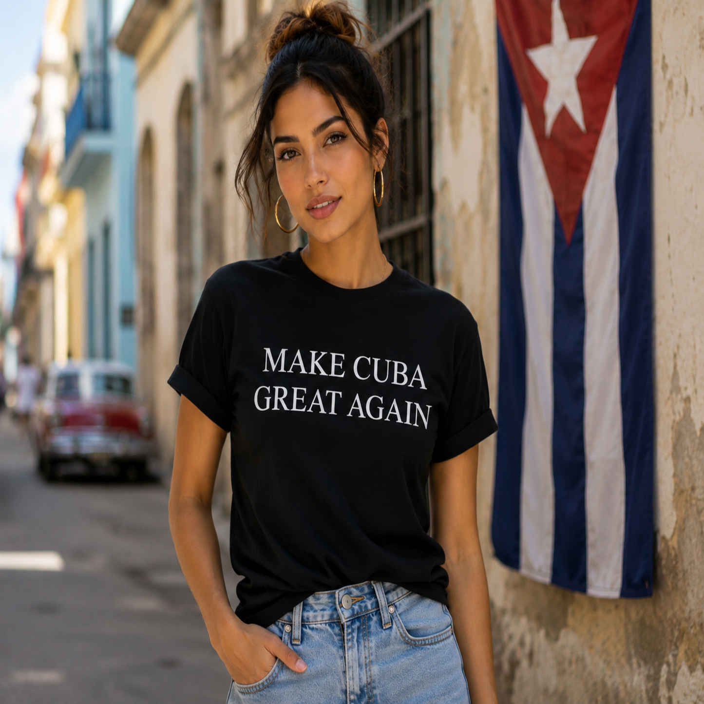 A woman wearing a black graphic t-shirt with the text 'MAKE CUBA GREAT AGAIN' in white capital letters, paired with light blue jeans, standing on a street next to a Cuban flag.